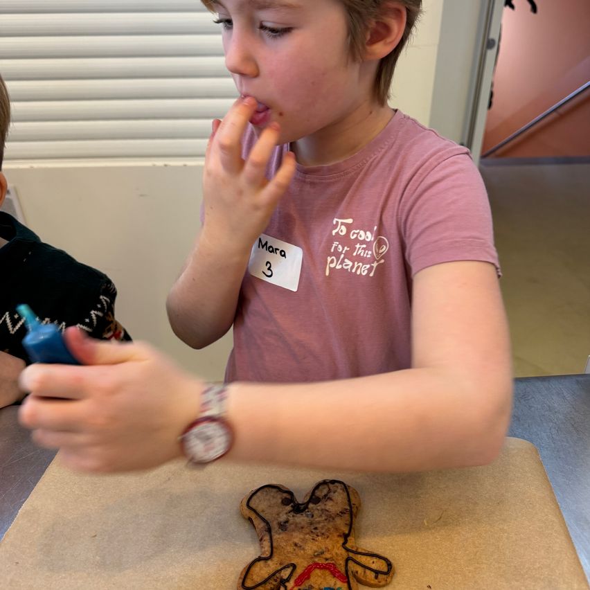 Ein Junge dekoriert einen Lebkuchenmann mit Zuckerguss. Er benutzt eine blaue Flasche mit Zuckerguss. Ein anderes Kind ist neben ihm. Sie befinden sich in einem Raum mit einer Treppe und einem Fenster.