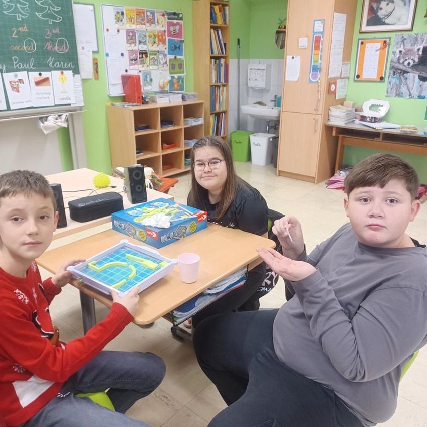 Drei Kinder sitzen an einem Tisch und spielen ein Brettspiel, wobei ein Mädchen lächelt und in die Kamera schaut.