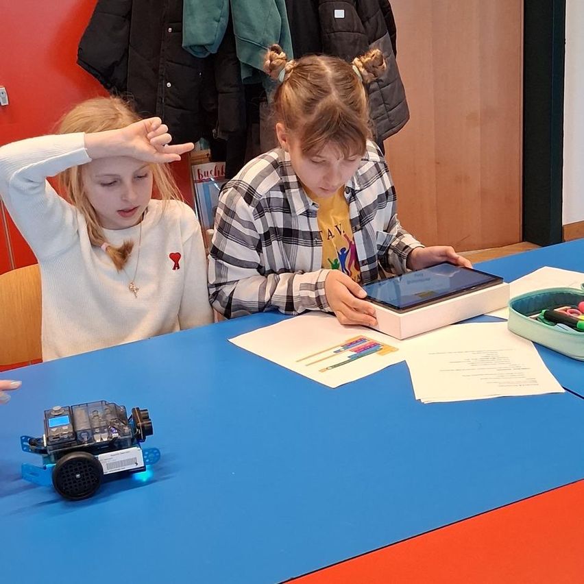 Zwei junge Mädchen sitzen an einem Tisch und beschäftigen sich mit einem Tablet. Eines der Mädchen schaut konzentriert auf den Bildschirm, während das andere gestikuliert. Ein Roboter und Papiere liegen auf dem Tisch. Kleidungsstücke hängen hinter ihnen.