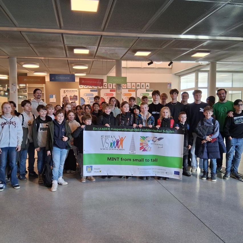 Eine Gruppe von Menschen, einschließlich Kindern, posiert für ein Foto in einem Schulkorridor mit einem Banner, das 'MINT von klein bis groß' liest.