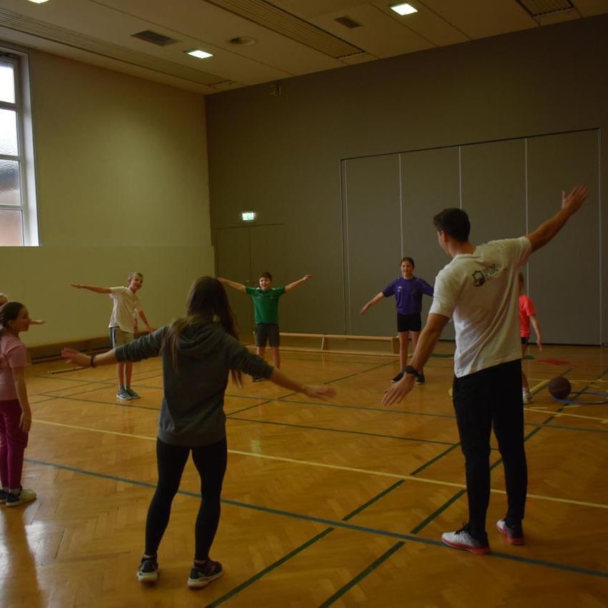 Eine Gruppe von Menschen trainiert in einer Turnhalle, ein Mann streckt seine Arme und andere beobachten. Der Boden ist aus Holz mit blauen Linien.