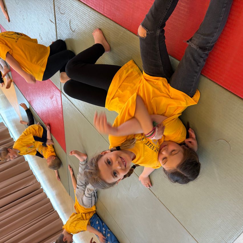 Eine Gruppe von Kindern in gelben T-Shirts übt Kampfsport auf Matten. Ein Mädchen liegt auf dem Boden, während zwei andere mit erhobenen Beinen auf dem Boden liegen.