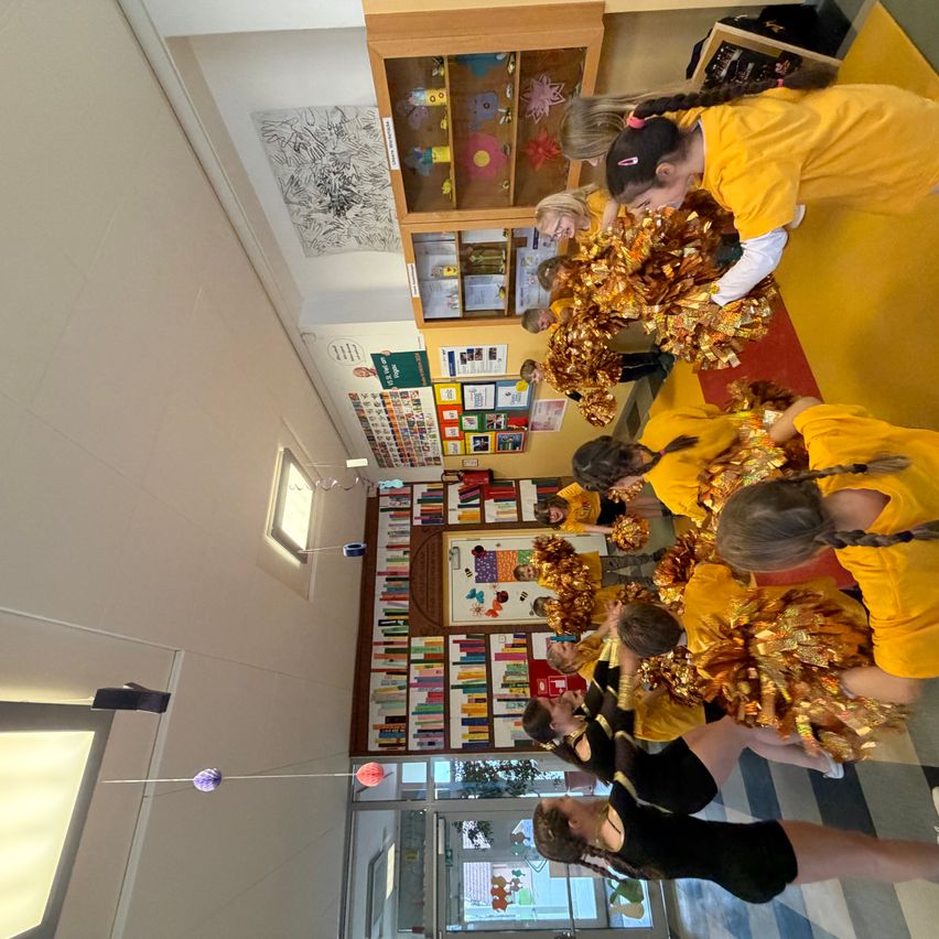 Eine Gruppe Cheerleader in gelben und schwarzen Uniformen steht in einem Flur, möglicherweise in Vorbereitung auf eine Veranstaltung. Der Raum ist mit Postern und Büchern an der Wand dekoriert.