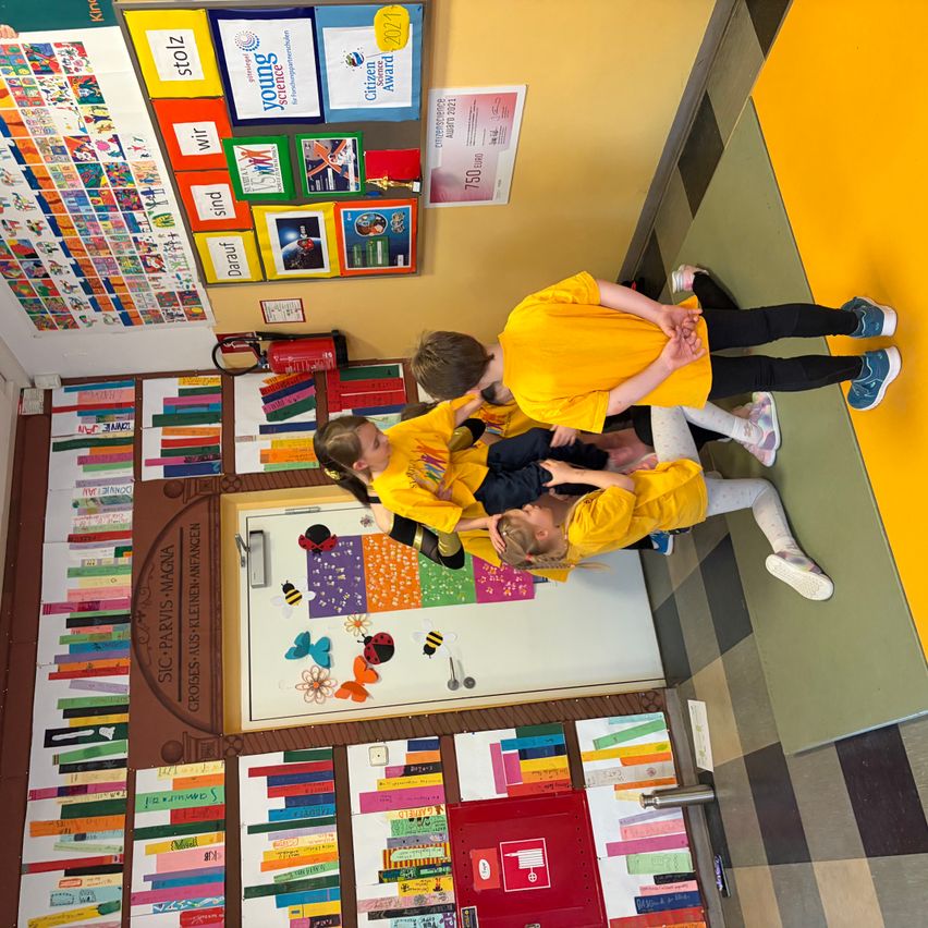 Vier Kinder spielen in einem Klassenzimmer. Drei von ihnen tragen gelbe T-Shirts. Sie stehen um eine Wand mit bunten Papieren.