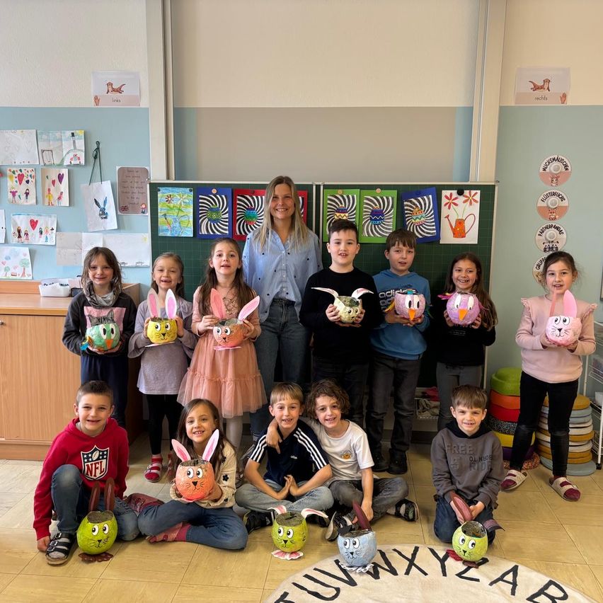 Eine Gruppe von Kindern mit ihren bemalten Osterhasen-Töpfen posiert für ein Foto mit einer erwachsenen Frau in einem Klassenzimmer.