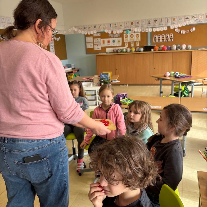 Eine Frau mit Brille steht in einem Klassenzimmer und hält ein Stück Essen. Mehrere kleine Kinder sitzen um sie herum und schauen zu.
