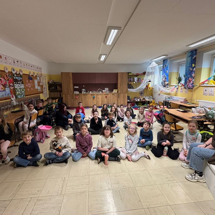Eine Gruppe von Kindern und Erwachsenen in einem Klassenzimmer, einige sitzen auf dem Boden, andere auf Stühlen, mit einem Holzschreibtisch, Schränken und Dekorationen im Hintergrund.