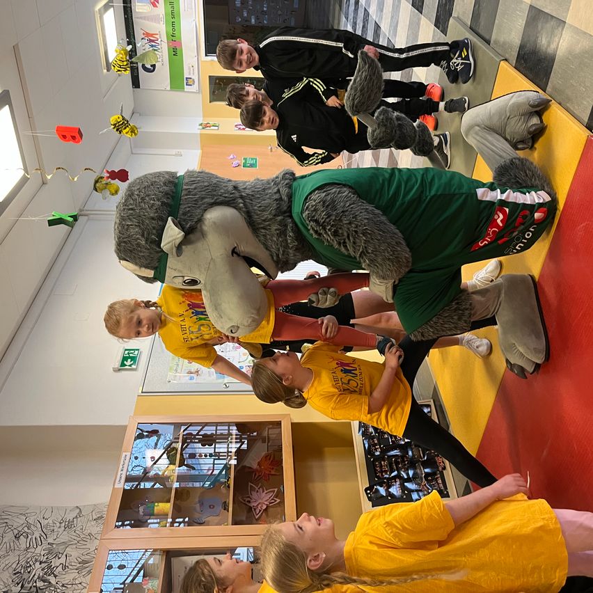 Mehrere Kinder in gelben Shirts und schwarzer Hose stehen mit einem Maskottchen. Das Maskottchen ist in einem grünen Shirt mit Logo gekleidet und hat einen grauen Körper. Einige Kinder schauen zum Maskottchen hoch. Es hängen Dekorationen von der Decke.
