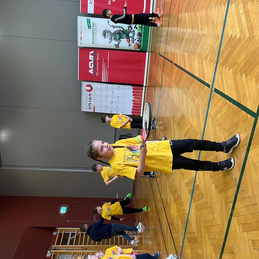 Mehrere Kinder in gelben Shirts spielen Badminton in einer Halle. Ein Kind hält einen Schläger und bereitet sich darauf vor, den Federball zu schlagen. Dahinter sind mehrere Plakate an der Wand angebracht.