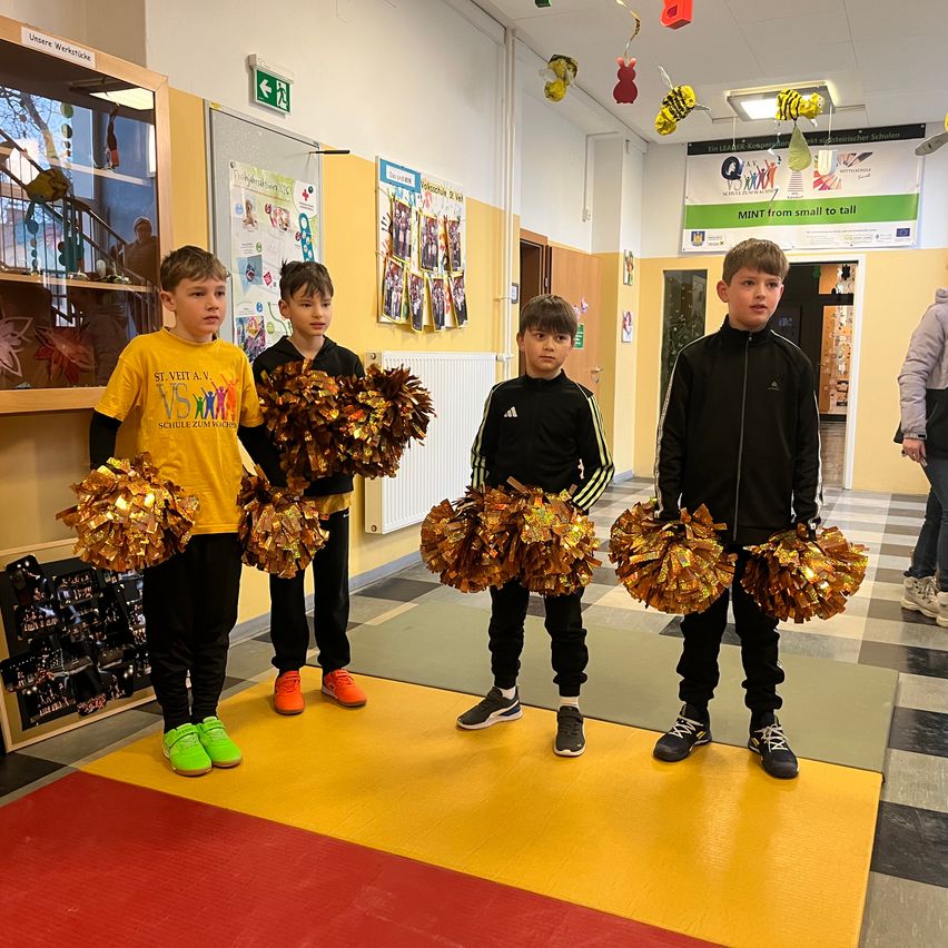 Vier junge Jungen stehen in einem Flur, jeder hält eine goldene Pom-Pom. Der Junge auf der linken Seite trägt ein gelbes Shirt mit VS darauf, während die anderen schwarze Jacken tragen. Hinter ihnen steht ein Glasschrank voller verschiedener Gegenstände.