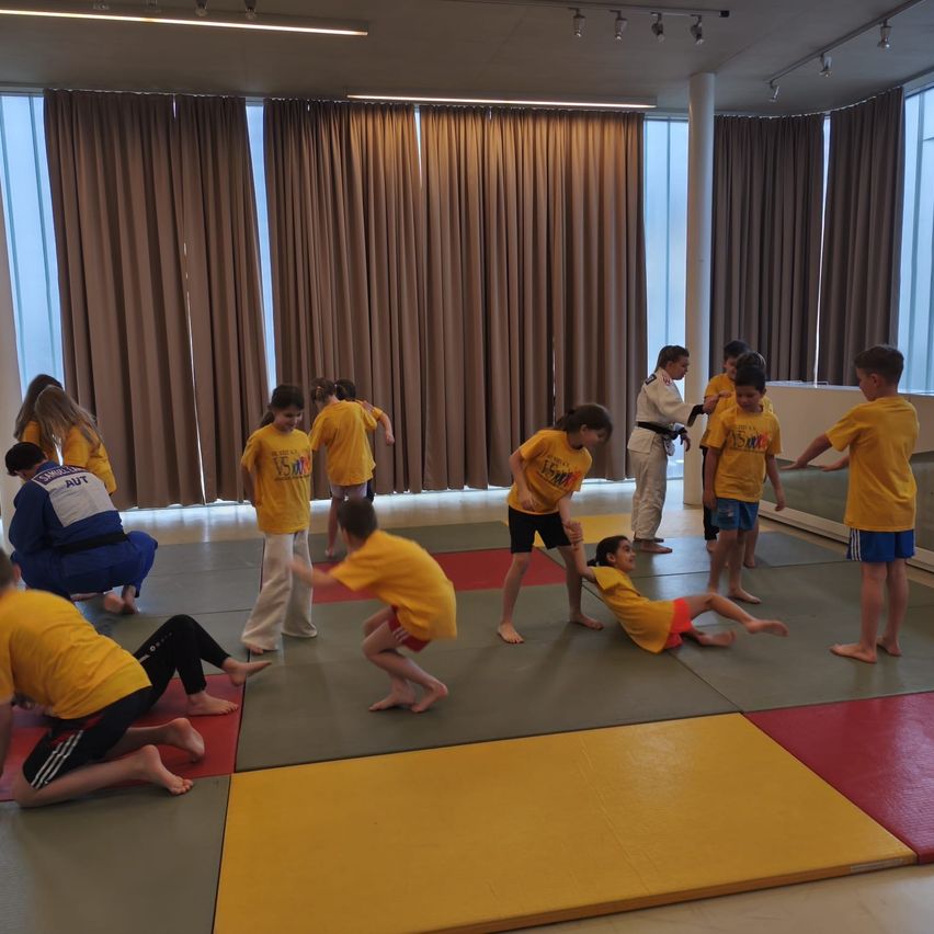 Mehrere Kinder in gelben T-Shirts üben in einem Raum mit Matten Kampfsport, während eine Person in einem blauen Gi zuschaut.