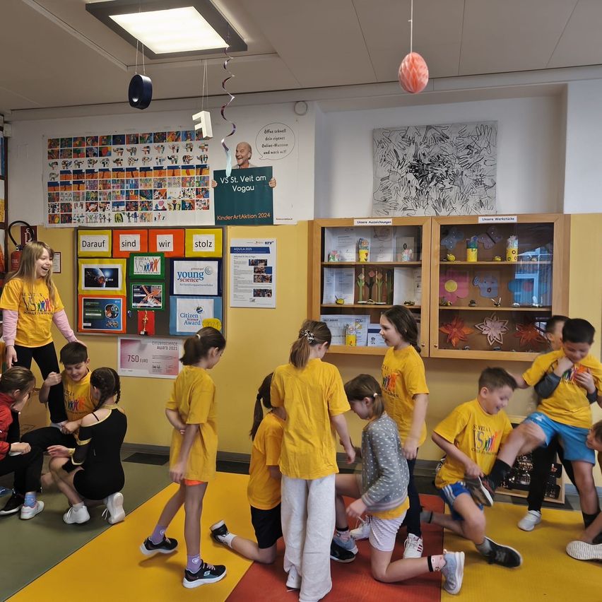 Eine Gruppe von Kindern in gelben T-Shirts führt eine Tanzroutine auf einem gelben Teppich in einem Klassenzimmer mit gelben Wänden und Regalen auf.