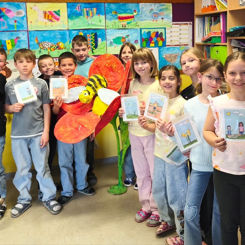 Eine Gruppe von Kindern steht vor einer Wand mit verschiedenen farbenfrohen Gemälden. Sie halten kleine Karten mit Bildern und einer bienenähnlichen Skulptur. Sie lächeln und scheinen für ein Foto zu posieren.