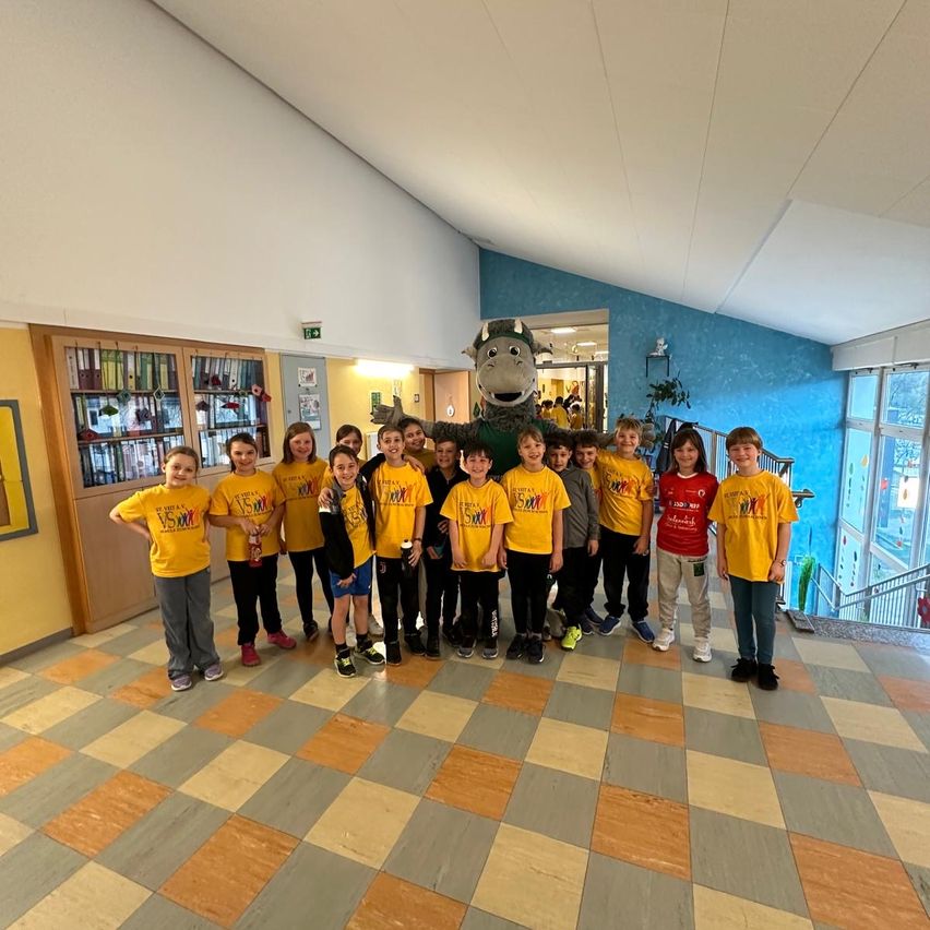 Eine Gruppe von Kindern in gelben T-Shirts posiert mit einem Maskottchen für ein Foto in einem Gebäude mit einem karierten Boden.