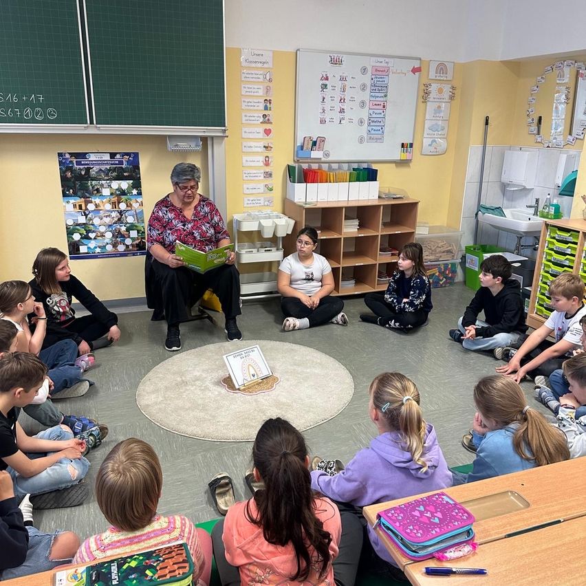 Eine Klassenzimmerszene, in der eine Lehrerin einem Kreis von Kindern, die auf dem Boden sitzen, ein Buch vorliest. Im Hintergrund sind verschiedene Lernmaterialien zu sehen.