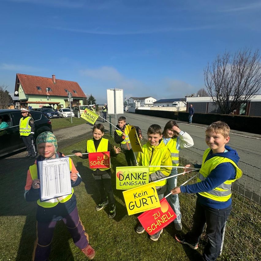 Eine Gruppe von Kindern in gelben Westen hält an einem sonnigen Tag Schilder. Sie stehen in einer Reihe und halten Schilder mit der Aufschrift 'Danke für Ihre Geduld!' und 'Keine Eile!'. Dahinter sind ein Haus und einige Autos zu sehen.