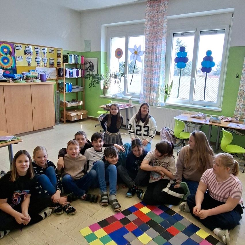 Ein Klassenzimmer mit Schülern auf dem Boden sitzend, einem bunten Quadrat auf dem Boden und einer stehenden Lehrerin. Grüne Wände und Fenster mit Luftballons.
