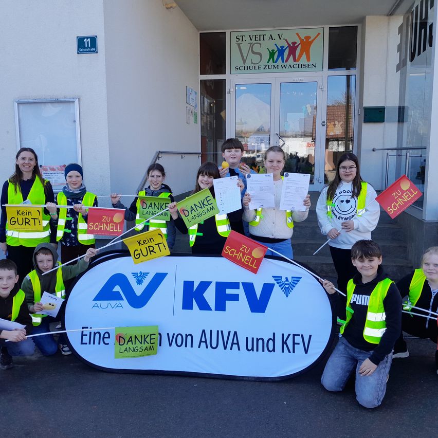 Eine Gruppe von Kindern in gelben Warnwesten mit Schildern posiert vor einem Banner mit der Aufschrift 'AV KFV'. Dahinter steht ein Gebäude mit einem Schild, das 'St. Veit a.v. VS' besagt.