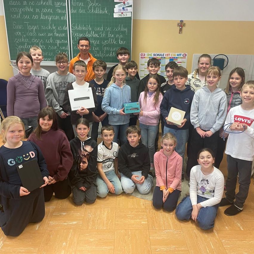 Eine Gruppe von Kindern und Erwachsenen posiert für ein Foto in einem Klassenzimmer. Ein Mann steht vorne und hält eine Box. Dahinter zeigt eine Tafel deutschen Text.