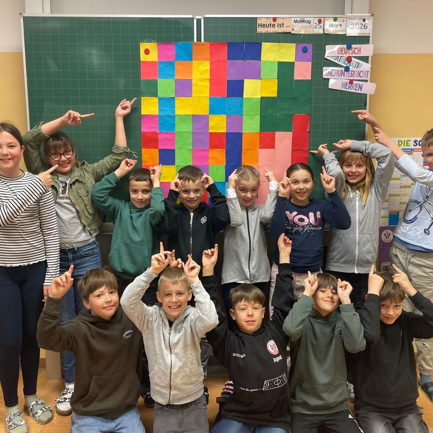 Gruppe von Kindern und Jugendlichen posiert für ein Foto in einem Klassenzimmer mit einem bunten Mosaik auf einer grünen Tafel.