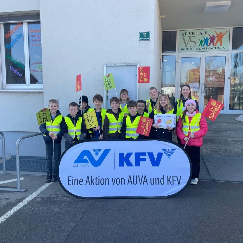 Eine Gruppe von Kindern in gelben Westen, die Schilder halten, steht vor einem Gebäude mit dem Schild AUVA und KFV. Dahinter befinden sich Glastüren und ein Gebäude mit dem Schild VS.