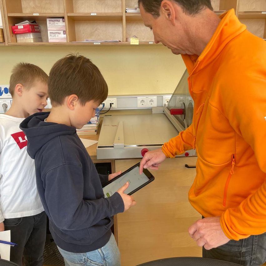 Ein Mann in einer orangefarbenen Kapuzenjacke instruiert zwei Jungen in einem Klassenzimmer, einer hält ein Tablet.