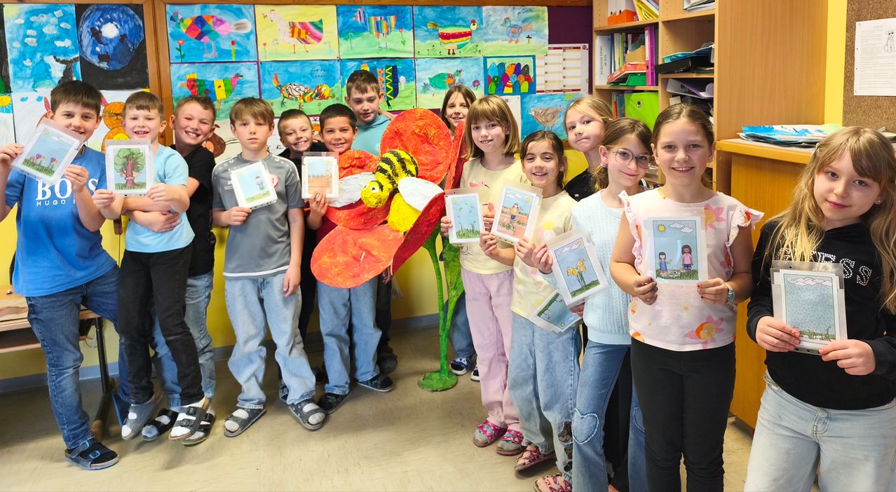 Eine Gruppe von Kindern posiert in einem Klassenzimmer, lächelt und hält Kunstwerke in den Händen. Eine bienenthematische Dekoration steht in der Mitte. Bunte Gemälde schmücken die Wände hinter ihnen.