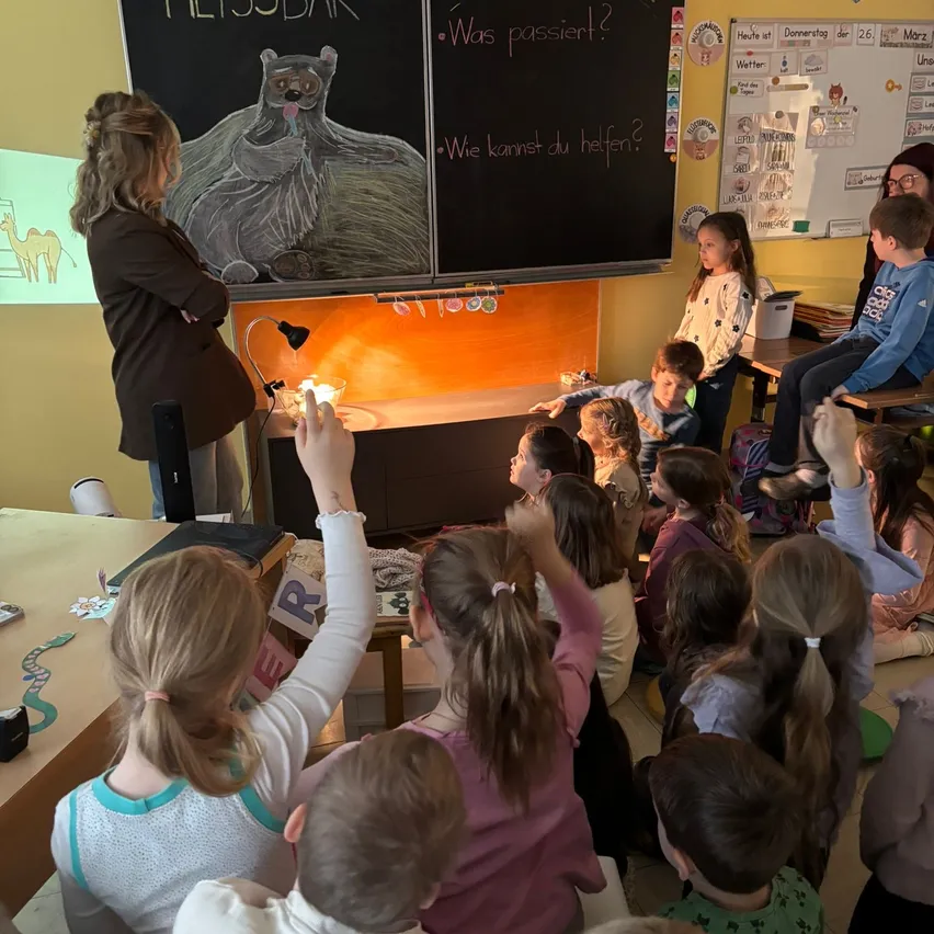 Eine Lehrerin steht an einer Tafel mit einer Bärenzeichnung. Kinder sitzen und stehen herum, einige mit erhobenen Händen. Ein Schreibtisch mit einer Lampe steht davor.