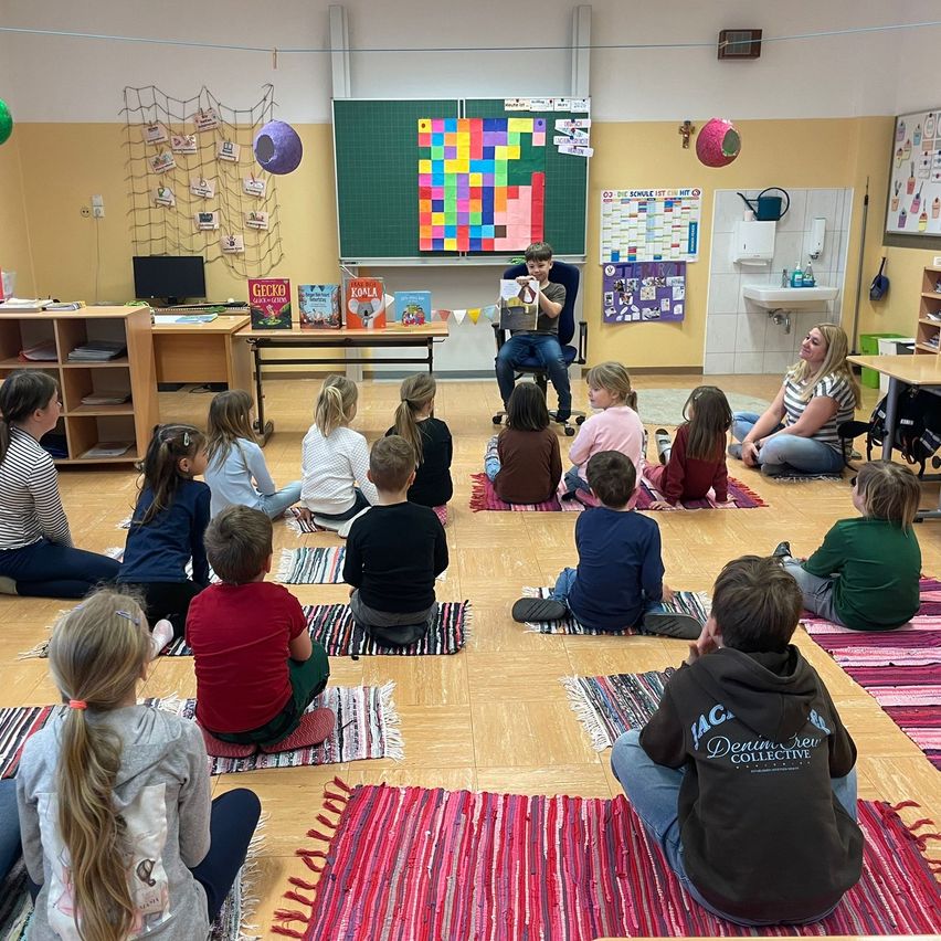 Eine Gruppe von Kindern sitzt auf dem Boden in einem Klassenzimmer. Ein Mann sitzt in einem Stuhl und liest ein Buch. Es gibt Bücher auf einem Schreibtisch in der Ecke.