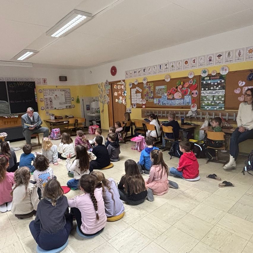 Ein Klassenzimmer mit Kindern auf dem Boden sitzend, einem Mann, der ein Buch liest, und einer Lehrerin, die mit den Schülern sitzt. An der Wand befinden sich Pinnwände mit verschiedenen Bildern und Wörtern.