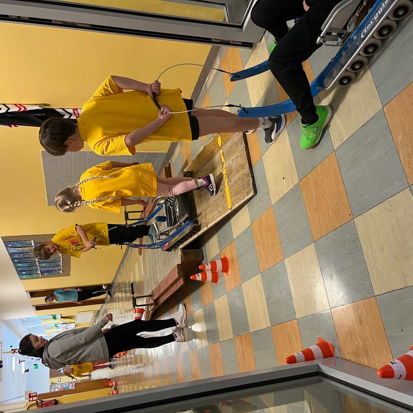 Eine Gruppe von Kindern in gelben T-Shirts nimmt an einer Schulaktivität teil, bei der ein Wagen und Kegel auf einem gefliesten Boden verwendet werden.
