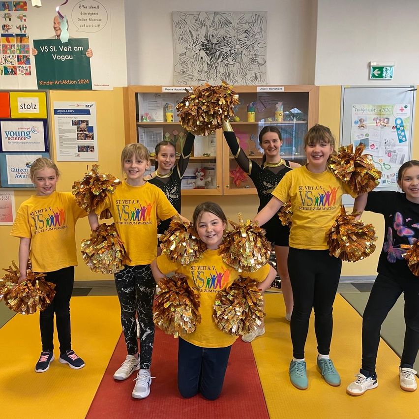 Gruppe junger Mädchen, die gelbe T-Shirts mit VS St. Veit am Vogau tragen, Pom-Poms halten und in einem gelben Raum mit Regalen und Postern an der Wand posieren.