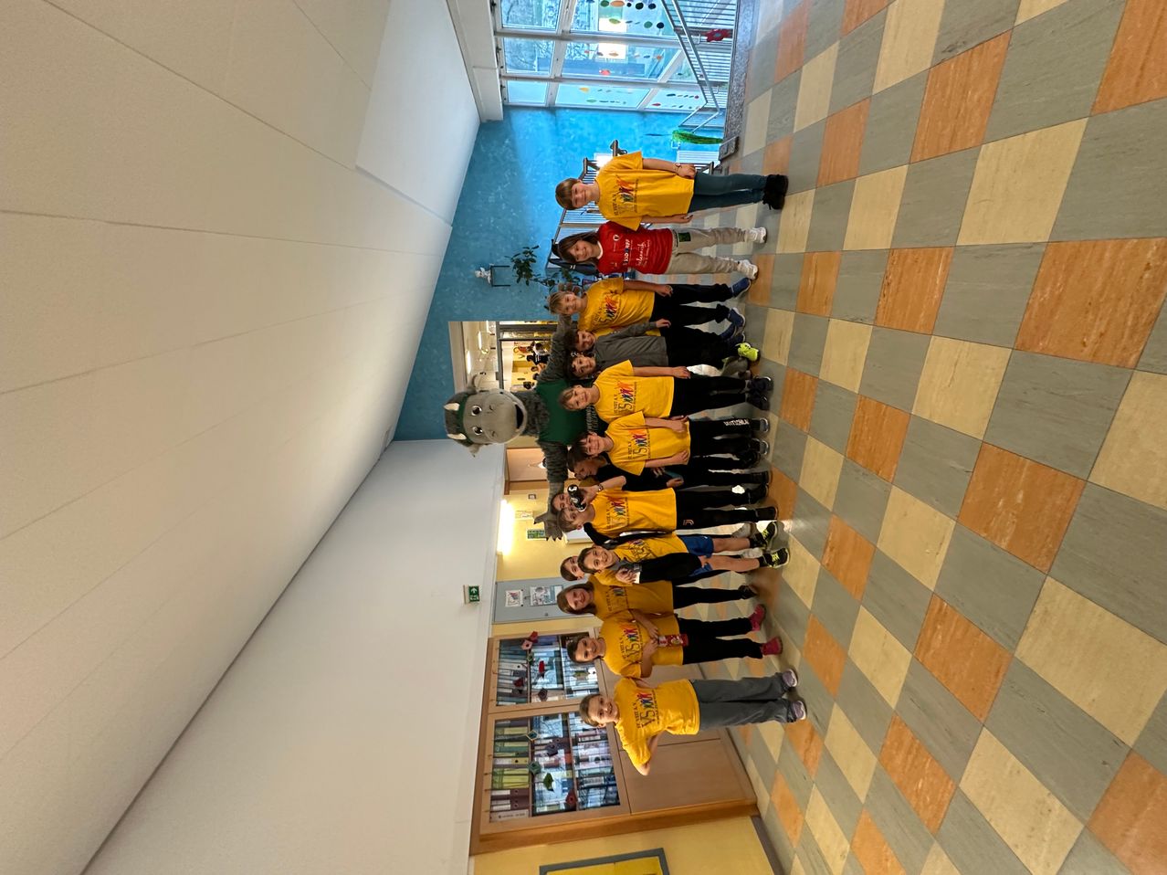 Eine Gruppe von Kindern und einem Erwachsenen, wahrscheinlich Lehrern, in gelben T-Shirts steht in einem Flur, mit einem Maskottchen in der Mitte. Der Flur hat einen karierten Boden und weiße Wände.