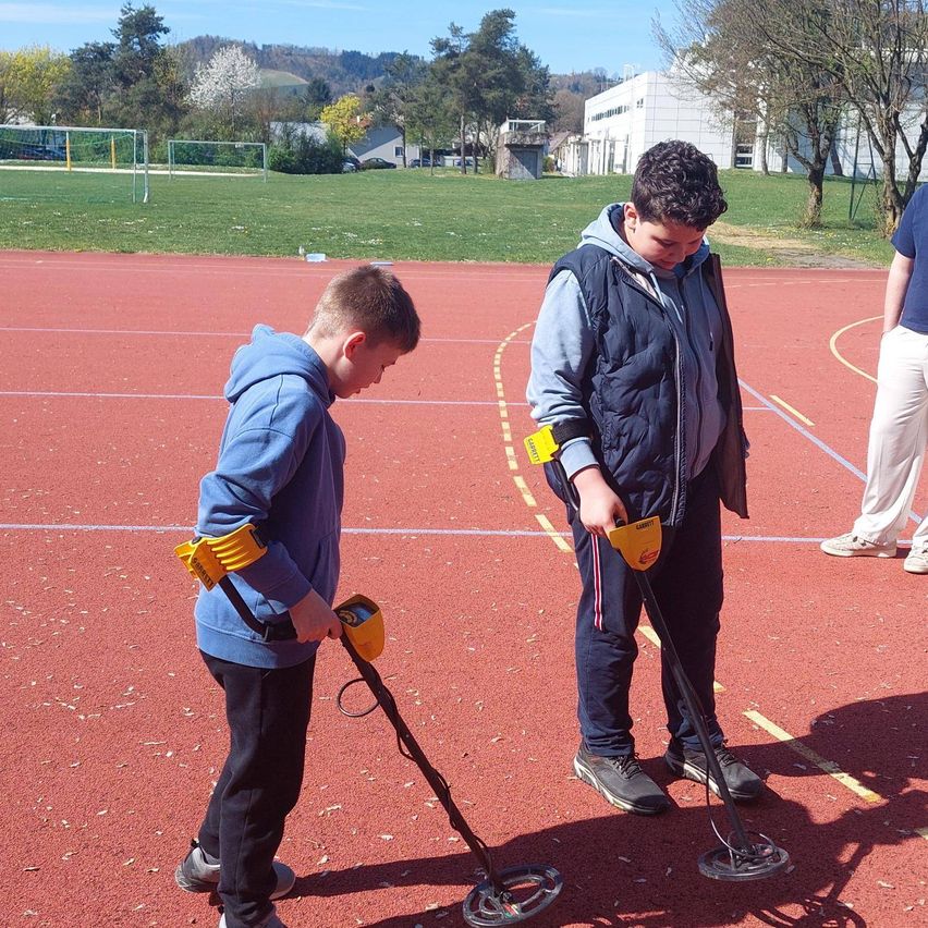 Zwei Jungen benutzen Metalldetektoren auf einer roten Laufbahn mit einem Grasfeld und Gebäuden im Hintergrund.