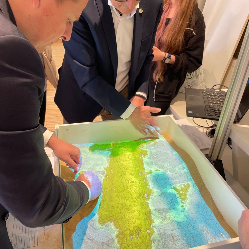 Mehrere Menschen in Anzügen untersuchen eine detaillierte Karte einer Insel, die auf einem Tisch projiziert wird, eine Person zeigt auf bestimmte Orte.