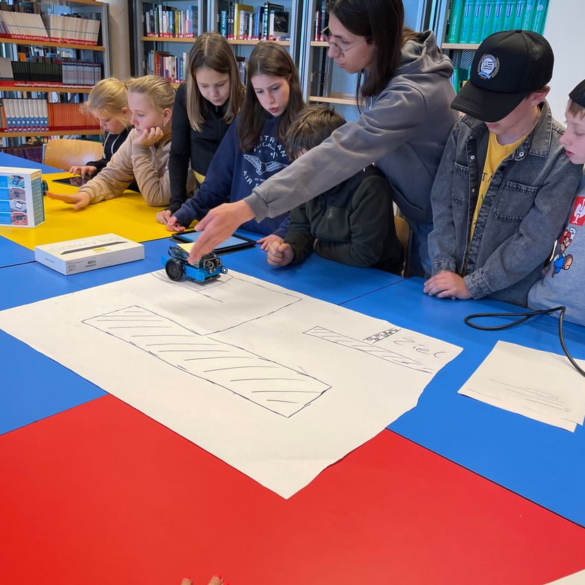 Eine Gruppe von Kindern und einem Erwachsenen versammelt sich um einen Tisch in einem Klassenzimmer. Sie arbeiten an einem blau-roten Tisch mit einem weißen Papier und einem blauen Spielzeugauto.
