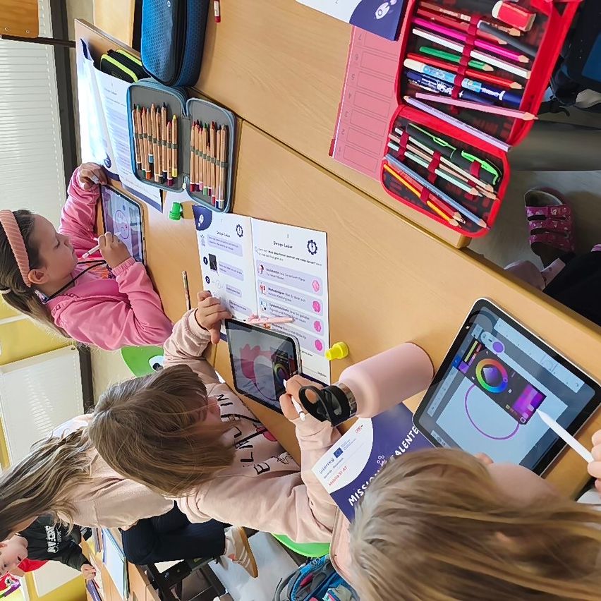 Mehrere Kinder sitzen um einen Tisch herum und benutzen Tablets. Ein Mädchen hält ein Tablet mit einer farbenfrohen Zeichnung. Der Tisch ist mit verschiedenen Zeichenmaterialien gefüllt.