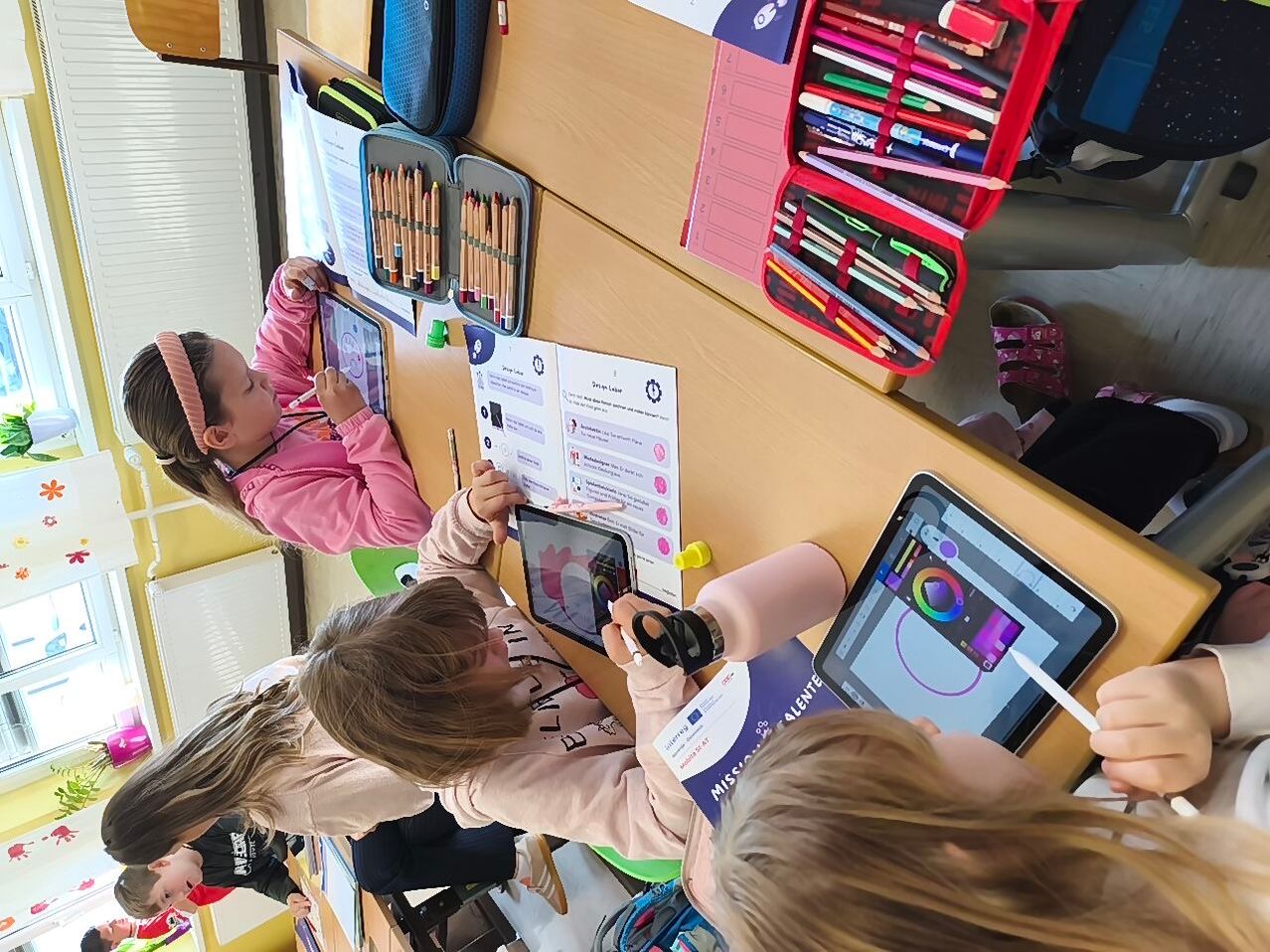 Mehrere Kinder sitzen um einen Tisch herum und benutzen Tablets. Ein Mädchen hält ein Tablet mit einer farbenfrohen Zeichnung. Der Tisch ist mit verschiedenen Zeichenmaterialien gefüllt.