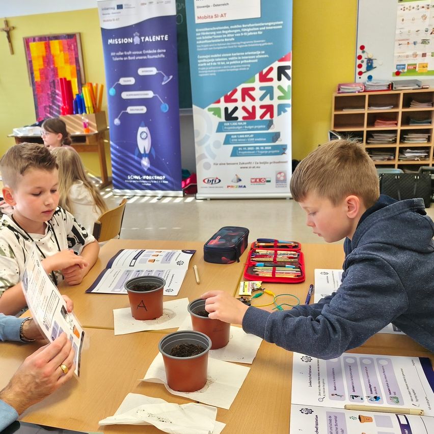 Zwei Jungen in einem Klassenzimmer pflanzen Samen in Töpfe, angeleitet von einem Erwachsenen. Andere Kinder sitzen hinter ihnen an einem Tisch.
