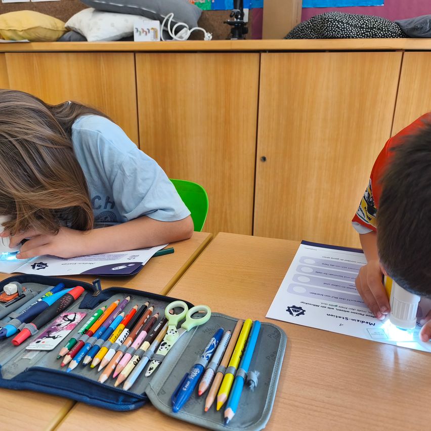 Zwei Kinder in einem Klassenzimmer benutzen eine Lupe, um ein Arbeitsblatt und eine Stifttasche voller farbiger Bleistifte und Kugelschreiber zu betrachten.