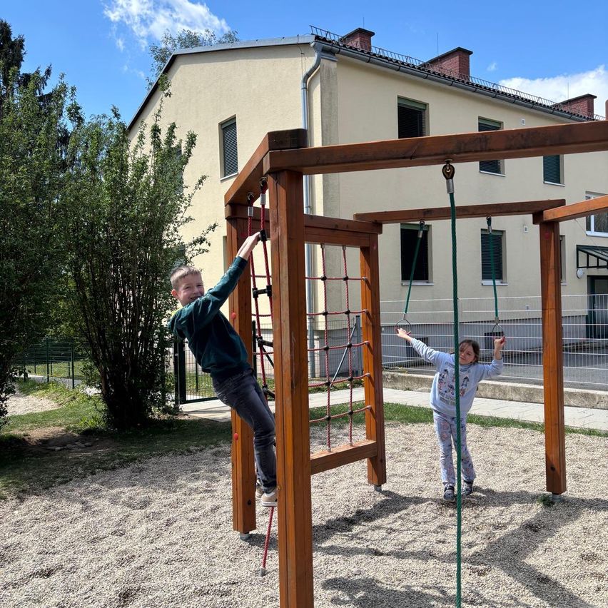 Zwei Kinder spielen auf einem hölzernen Klettergerüst in einem Park. Ein Junge klettert eine rote Seilleiter hinauf, während ein Mädchen eine Schaukel hält. Dahinter steht ein beiger Haus mit Fenstern und einem Schornstein.