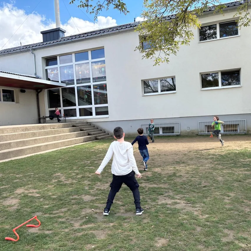 Mehrere Kinder spielen vor einem Gebäude mit großen Fenstern und Treppen. Ein Kind läuft, andere stehen still. Ein rotes Objekt liegt auf dem Gras.