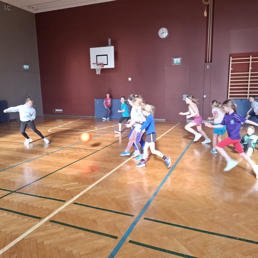 Kinder spielen eine Partie Dodgeball auf einem Hallensportplatz. Ein Kind wirft einen Ball. Der Platz hat Holzboden und blaue Markierungen.