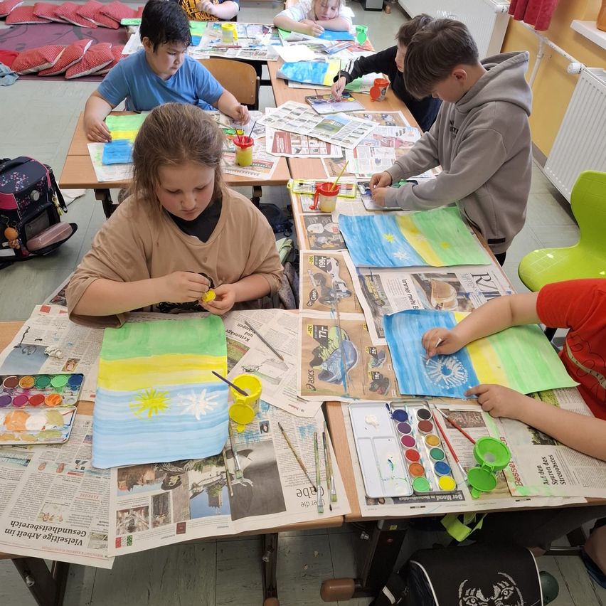Schüler in einem Klassenzimmer malen auf großen Papierbögen. Einige konzentrieren sich auf ihre Kunstwerke, während andere Wasserfarben verwenden.