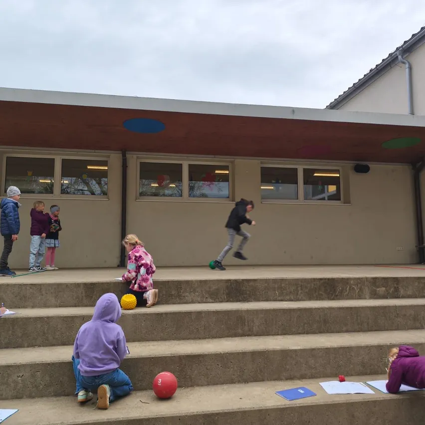 Kinder auf einer Schultreppe spielen Ballspiele. Ein Junge springt mit einem Ball in der Hand. Zwei Kinder schreiben auf Papier.