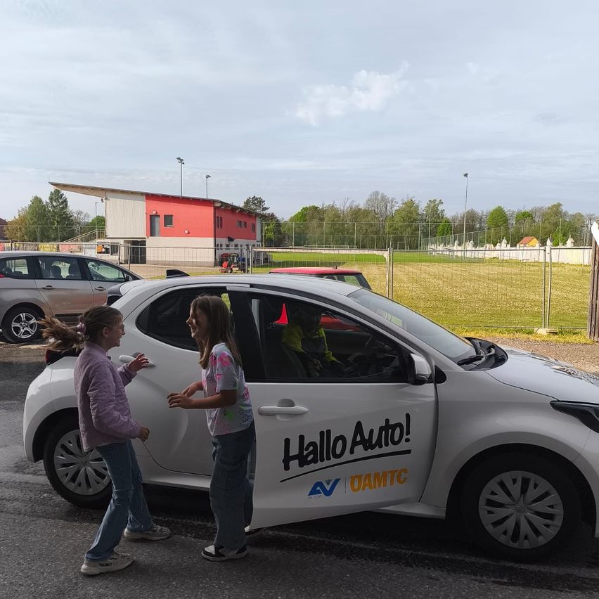 Zwei Mädchen stehen vor einem weißen Auto mit dem Text Hallo Auto! darauf. Ein Mann sitzt im Auto. Hinter dem Auto steht ein Gebäude, und ein Maschinenfahrzeug ist davor geparkt.