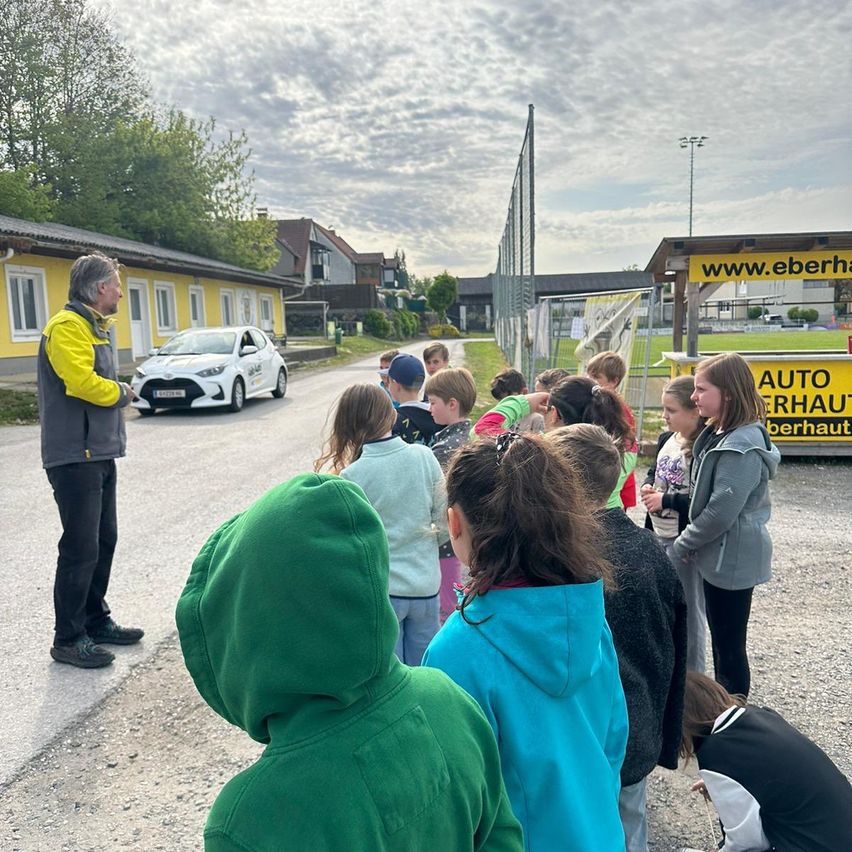 Ein Mann in einer gelben und grauen Jacke spricht mit einer Gruppe von Kindern bei einem Auto auf einer Straße.