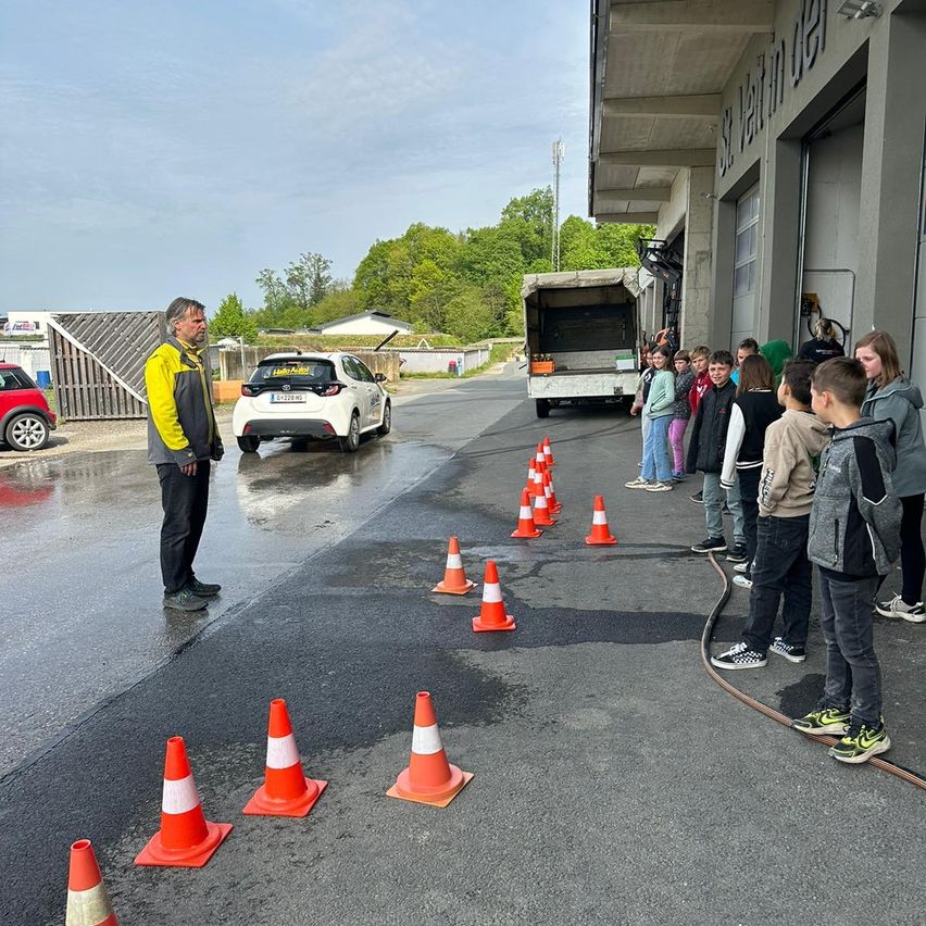 Ein Mann steht in einem nassen Bereich mit Verkehrskegel und Kindern in einer Reihe. Ein Lkw und ein Auto sind geparkt.