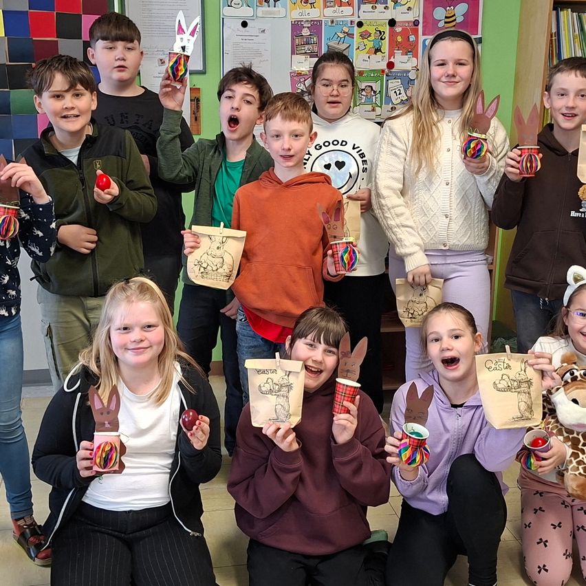 Eine Gruppe von Kindern in einem Klassenzimmer hält dekorierte Osterkörbe und trägt Hasenohr-Kopfbänder. Sie posieren für ein Foto.