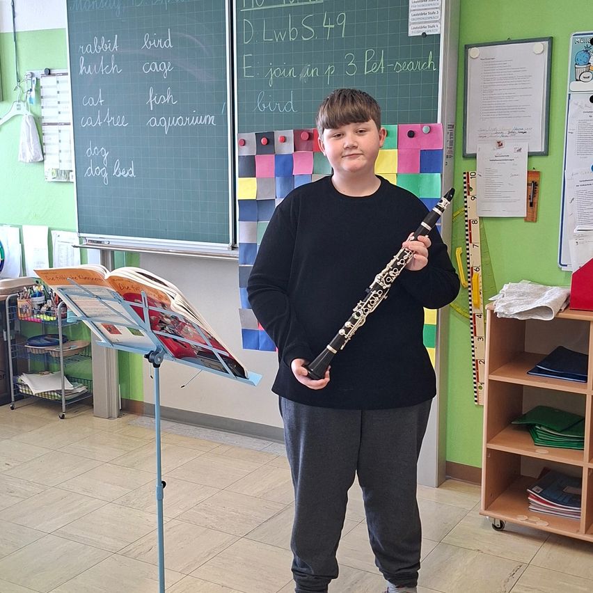 Ein junger Junge steht in einem Klassenzimmer mit einer Klarinette in der Hand, mit einem Notenständer und offenem Buch dahinter. Eine Tafel hat Wörter und Zahlen darauf geschrieben.